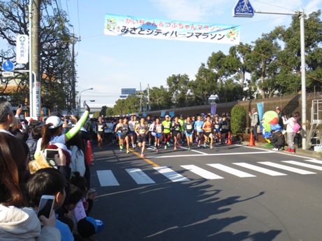 車道に設けられたマラソンコースで、頭上に掛かったスタート地点を示す「みさとシティハーフマラソン」の横断幕の下を走っていくたくさんの参加者たちと、沿道で応援をするたくさんの観客の写真