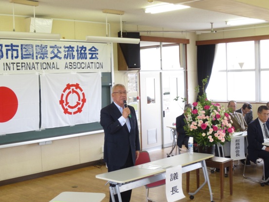 「三郷市国際交流協会」と書かれた横断幕と国旗・三郷市旗が黒板に貼り付けられ、その前に配置されたピンク・白・緑の花々で彩られた花台と並ぶ議長席のそばに立ちあいさつの言葉を述べる三郷市長と、席に座って話を聞く関係者の写真