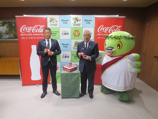 両側に「CocaCola」の文字と白いボトルのイラストが描かれた赤色のバナースクリーンが設置されている三郷市のバックボードの前で、市川朋弘コカ・コーラ ボトラーズジャパン株式会社ベンディング関東地区統括部地区統括部長と木津雅晟市長が聖火リレーデザインをラッピングした沢山の清涼飲料水が置かれている緑のカバーが掛けられた台を挟んで立ち、オリンピックの衣装を着用したかいちゃんとともに記念撮影をしている写真