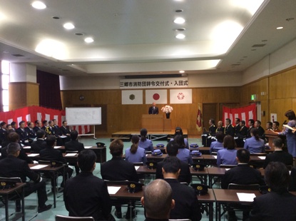 「三郷市消防団辞令交付式・入団式」と書かれた横断幕の下の檀上に立ち挨拶を行っている市長の話を着席して聞いている団員の後ろ姿の写真