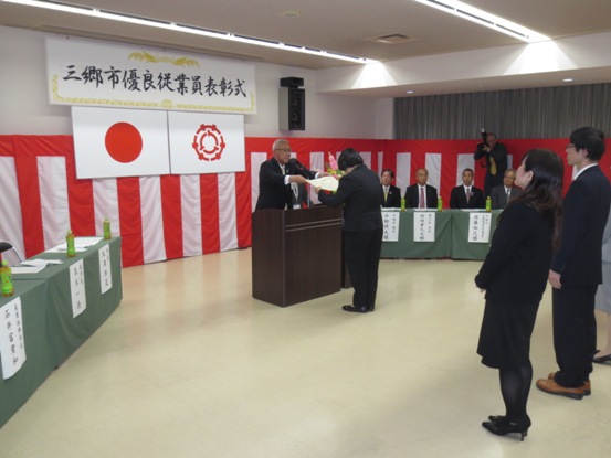 「三郷市優良従業員表彰式」と書かれたボードと国旗・三郷市旗が吊り下げられている壁の前で、演台に立ち表彰状を手渡す三郷市長と向かい合って立ち両手で受け取る女性、後ろに横一列に並ぶ複数名の優良従業員の写真