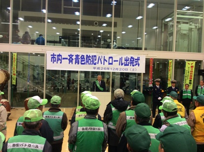 「市内一斉青色防犯パトロール出発式」と書かれた横断幕の下に設置された檀上に緑色のタスキを肩にかけた市長が立ち挨拶を行っているのを防犯パトロールと書かれた緑色のゼッケンを着用した参加者が聞いている後ろ姿の写真