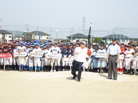 半田運動公園のグラウンドで、左手にグローブをはめて右手で野球ボールを投げる三郷市長と、前列の子どもがチーム名の入った立て看板を持ちチームごとに整列している子どもたちの写真