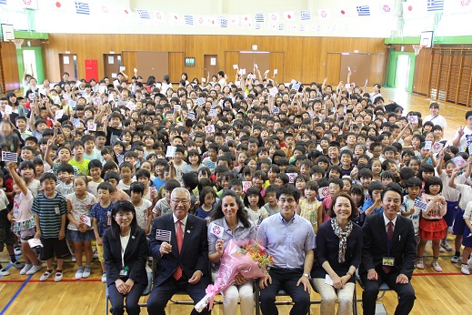 最前列に三郷市長と花束を持ったギリシャ共和国大使夫人、大使館職員が座り、後方にギリシャの国旗と三郷市旗を持った大勢の子どもたちが集まり記念撮影をしている写真