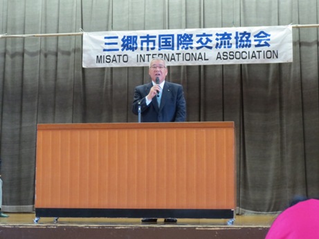「三郷市国際交流協会」と書かれた横断幕の下の演壇で、右手にマイクを持った市長が挨拶を行っている写真
