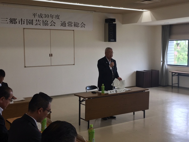 壁に「平成30年度三郷市園芸協会通常総会」の紙が貼ってある部屋で、正面中央に設置された長机の席から起立してマイクを手に話をしている市長の写真