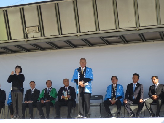 産業フェスタ式典で挨拶の言葉を述べる青い法被を着用した三郷市長と近くに立ち手話通訳をする女性、後ろの壁沿いに並べられたパイプ椅子に座り話を聞く関係者の写真