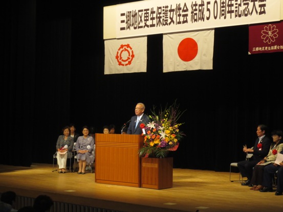 「三郷地区更生保護女性会結成50周年記念式典」のボードが吊られた舞台上で、演壇に立って話をしている市長の写真
