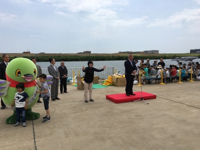 右側で奥に流れる川の方を向いて座っている方々、左側にマスコットキャラクターかいちゃんの傍から離れない2名の男の子、中央に黒色のシャツを着ている女性の手話と同時に小さな台の上に立っている市長が挨拶を行っている写真