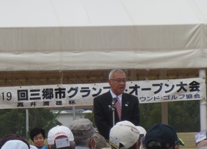 「第19回グランプリ・オープン大会高井満雄杯」と書かれた横断幕の前に立ち挨拶の言葉を述べる三郷市長と、市長の方を向いて話を聞く、名札のついた帽子をかぶった参加者の写真