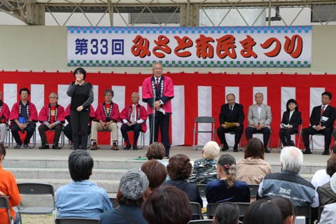 「第33回みさと市民まつり」と書かれた横断幕と紅白幕が貼られた屋外ステージに設置された椅子に関係者が座り、手話通訳者の女性と挨拶をしている市長が中央に立っている写真