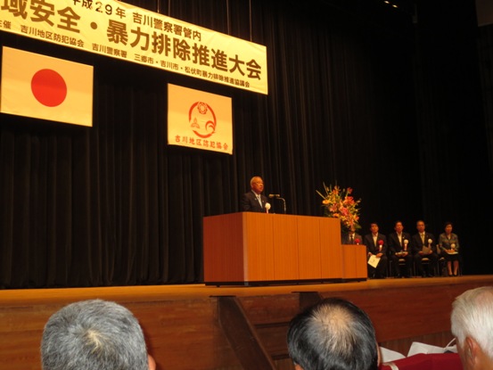 「地域安全・暴力排除推進大会」と書かれた横断幕と国旗・吉川地区防犯協会の旗がステージ上に吊るしてあり、豪華な花々で彩られた花台と並ぶ演台に立ち挨拶の言葉を述べる三郷市長と、ステージ上のパイプ椅子に座る胸章を付けた来賓者の写真