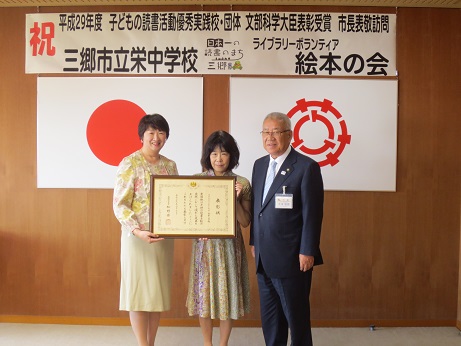 「祝平成29年度子どもの読書活動優秀実践校・団体文部科学大臣表彰受賞市長表敬訪問」と書かれたポスターと国旗・三郷市旗が吊るされた壁の前で、ライブラリーボランティア絵本の会のメンバー2名が表彰状を一緒に持ち三郷市長と並んで記念撮影をしている写真