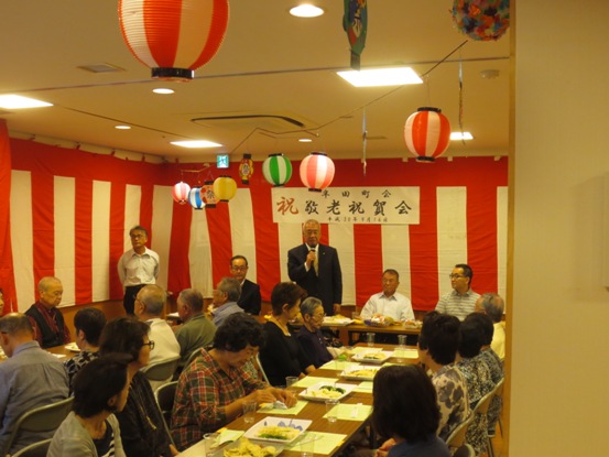 紅白の横断幕や提灯で飾られている部屋で、料理が並んだ長机に着席にしている参加者と、正面に立って話をしている市長の祝賀会の様子の写真