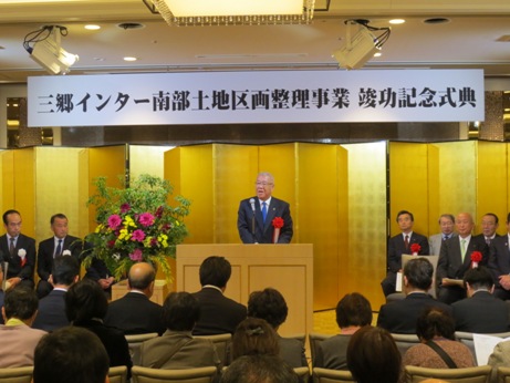 「三郷インター南部土地区画整理事業竣功記念式典」と書かれた横断幕と金屏風の前の演壇で挨拶を行っている市長の写真