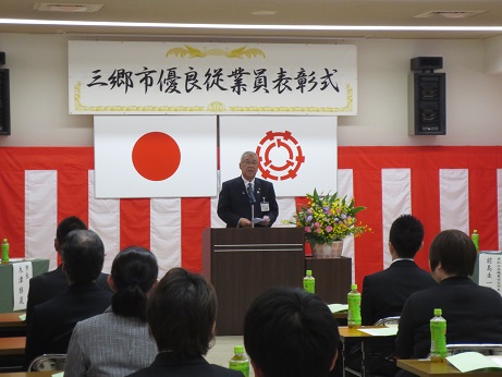 「三郷市優良従業員表彰式」と書かれたボードと国旗・三郷市旗が天井から吊り下げられ、壁に紅白幕が飾られた会場で、ピンク・黄色・紫の花々で彩られた花台と並ぶ演台に立ち挨拶の言葉を述べる三郷市長と、ペットボトルのお茶が置かれている席に座り話を聞く受賞者の写真