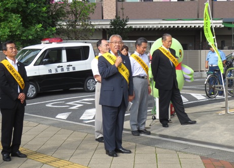 後方の駐車場にパトカーが駐車している手前にマスコットキャラクターかいちゃんと黄色のタスキを肩にかけた4名の関係者が横一列に並び、一歩前に右手にマイクを持った市長が挨拶を行っている写真