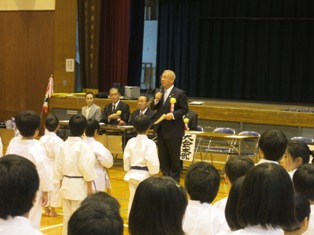 「大会本部」と書かれた長机の前に立ち挨拶を行っている市長の話を道着を着て並んで立っている子供たちが聞いている写真