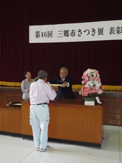 三郷市長賞の作品が置かれた演台の前に立って、市長から表彰を受けている男性の写真