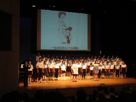 「社会を明るくする運動」子どもが母親に抱きついているイラストがスクリーンに映し出され、水色のスカーフを首に巻いた大勢の前谷小の児童たちが舞台に上がり合唱をしている写真