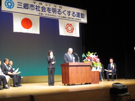 「三郷市社会を明るくする運動」と書かれたプレートと国旗、三郷市章が掲げられた会場で市長が演台に立ち式辞を述べ、左脇に女性が手話通訳をし、関係者の方々がパイプ椅子に座って聞いている写真