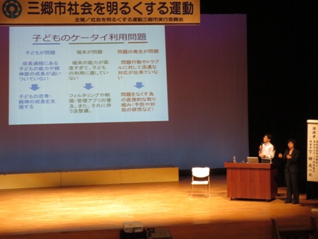 「三郷市社会を明るくする運動」と書かれたプレートが掲げられた会場で、講演内容の「子どものケータイ利用問題」をスクリーンに映し出し、演台に立ちマイクを持って講演している下田太一先生と、女性が手話通訳している講演会の様子の写真