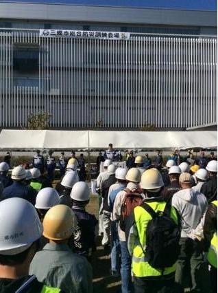 「三郷市総合防災訓練会場」と書かれた横幕が飾られ、テントの前の檀上に立つ市長の話をヘルメットを被っている参加者が聞いている後ろ姿の写真