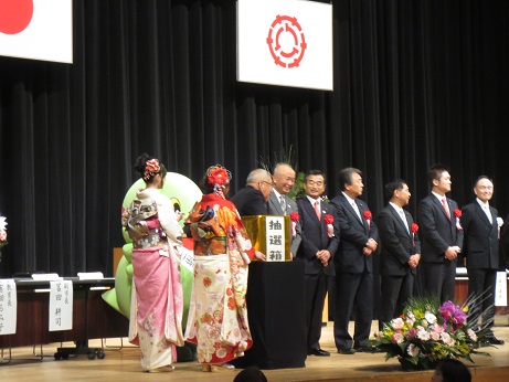日の丸と三郷市の旗が掲げられた舞台の上で、来賓が横一列に並んで立っており、その向かって左端にいる市長が抽選箱に手を入れているところを、舞台左側に立っている振袖姿の新成人2人とかいちゃん&つぶちゃんが見ている様子の写真