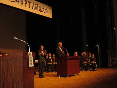 舞台中央に設置された演台に立ちマイクに向かって挨拶をする市長と、その向かって左後方に立ち手話通訳をする女性、右後方で2列に座った来賓を写した写真