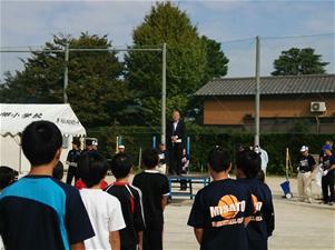 八木郷小学校の校庭の朝礼台に立ち挨拶を行っている市長の話を聞いている子供たちの後ろ姿の写真
