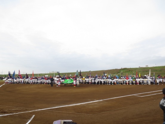 チームごとに整列した大勢の選手たちの最前列者がプラカードを持ち、列の中からは色とりどりのチーム旗が上がっており、選手たちの前で4名の選手が大きな緑の旗の四隅をそれぞれ持っている入場行進の様子の写真