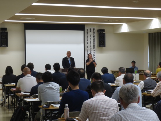 等間隔に並べられた長机に2脚ずつの椅子が用意され、椅子に着席している関係者たちと、正面の演壇に立って挨拶をしている市長の写真