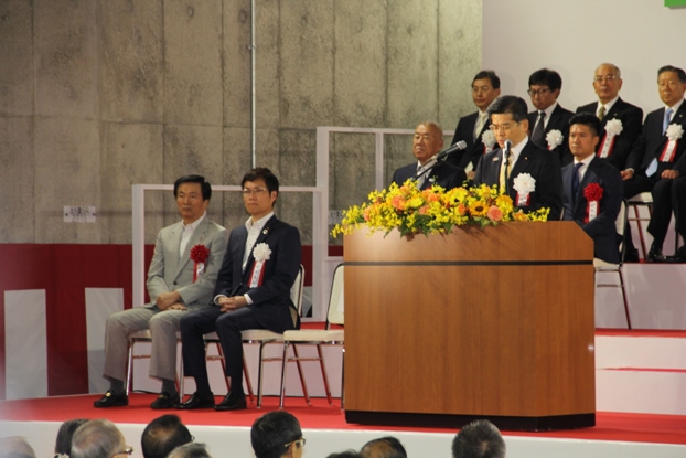 スーツ姿の関係者たちが胸章をつけ着席しているなか、石井国土交通大臣が演台に立って挨拶をしている写真