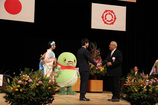 国旗と三郷市旗が吊るしてあり、ステージ前方と花台には色とりどりの豪華な式典花が飾られているステージ上で、演台の前で握手をする記念品を手に持った三郷市長と抽選会で当選した男性、その様子を見守る薄ピンクと水色の配色で花のデザインが施された着物を着用した女性と白いタスキをかけたかいちゃんの写真