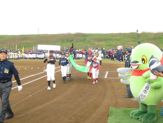 チームのプラカードを持った選手の後ろを4名の選手が緑の旗の四隅をそれぞれ持って歩いており、かいちゃんとつぶちゃんが周りに立つ関係者とともに選手を出迎えている写真