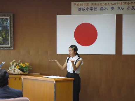 国旗が吊り下げられている壁の前の演台に立った、青少年の主張大会で最優秀賞を受賞した鈴木奏さんが、両手のひらを天井に向けたジェスチャーをしながら結果報告と発表を行っている様子の写真