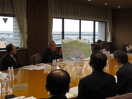 三郷市の広域図を見せながら三郷のまちづくりの状況を説明する三郷市長と、水と資料が置かれたテーブルを囲んで座り話を聞く関係者の写真