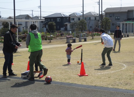 芝生の上に置かれたバッティングティーの横で、ピンクのジャンパーを着た小さな女の子が赤いバットを手に持ち、その様子を男性が見守っているティーボール体験の様子の写真