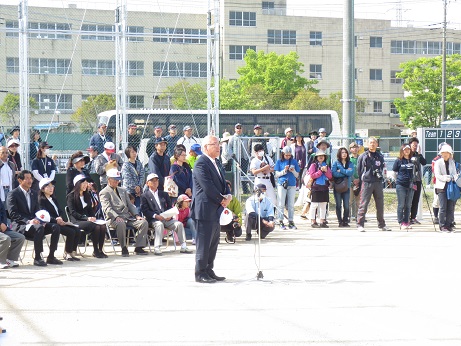半田運動公園のグラウンドに立ちあいさつの言葉を述べる三郷市長と、三郷市長の後ろに横に並び話を聞く関係者の写真