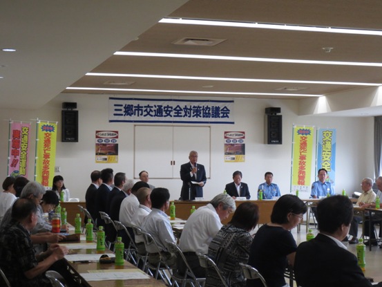 壁に「三郷市交通安全対策協議会」の横断幕が貼られた部屋で、資料が置かれた長机が並び、多くの関係者が椅子に着席し集まっているなか、正面に立つ市長が話をしている写真