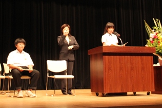ステージ上に設置された椅子に男子生徒が座り、右側の演壇に設置されたマイクの前でスーツ姿の女性の手話と同時に発表を行っている女子生徒の写真