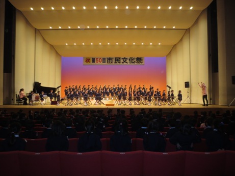 「祝第50回市民文化祭」と書かれた横断幕がステージ上に掛けてあり、ステージ右手に立つ指揮者を見ながら合唱をするわせだ幼稚園の大勢の子どもたちと、ステージ左手に置かれた黒いグランドピアノを演奏する女性、ドラムなどの楽器を演奏する子どもたちの写真