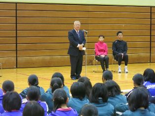 体育館の右側に神白氏と山本氏が椅子に座り、マイクスタンドの前に立ち挨拶を行っている市長の話を床に体育座りをして聞いている中学生の後ろ姿の写真