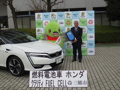 「燃料電池車 ホンダ クラリティ」と書かれた看板の後ろに駐車された白い自動車の横で、かいちゃんと、大きな車のレプリカキーを持った市長が三郷市のパネルの前に立っている写真