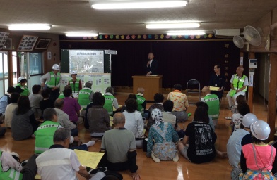 公民館に設置された檀上に立ち挨拶を行っている市長の話を床に座って聞いている参加者の後ろ姿の写真