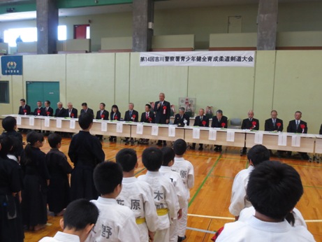 「第14回吉川警察署青少年健全育成柔道剣道大会」と書かれた横断幕が掲げられた壁を背に、横一列に並んだテーブルに着席しているたくさんの来賓の中央に立ち話をする市長と、来賓席を向いて整列した剣道や柔道の道着姿の子供たちの写真