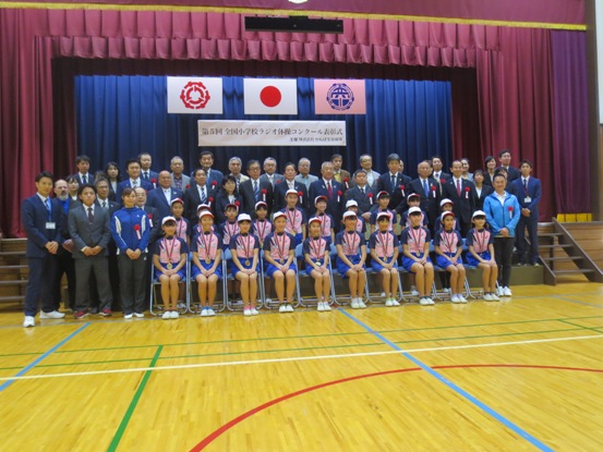 体育館でメダルを肩から下げた小学生が横2列に並び、その横や後ろにたくさんの関係者が一緒に並んで記念撮影をしている写真