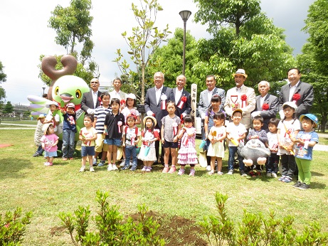 胸章を付けた三郷市長・関係者・子どもたちが、かいちゃん・つぶちゃんとともににおどり公園の広場で記念撮影をしている写真