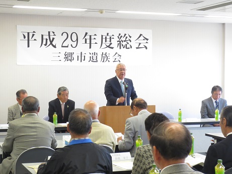 「平成29年度総会三郷市遺族会」と書かれているポスターが貼りつけられた壁の前で、演台に立って挨拶の言葉を述べる三郷市長とペットボトルのお茶が置かれた席に座り資料に目を通しながら話を聞く遺族の写真