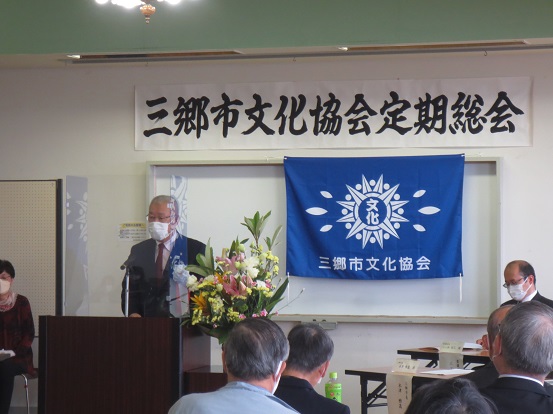 「三郷市文化協会定期総会」と書かれた横断幕と三郷市文化協会の青い旗が掲げられた会場で、三郷市長が演台に立って挨拶をし、関係者の方々と参加者の方々が席に座って聞いている写真